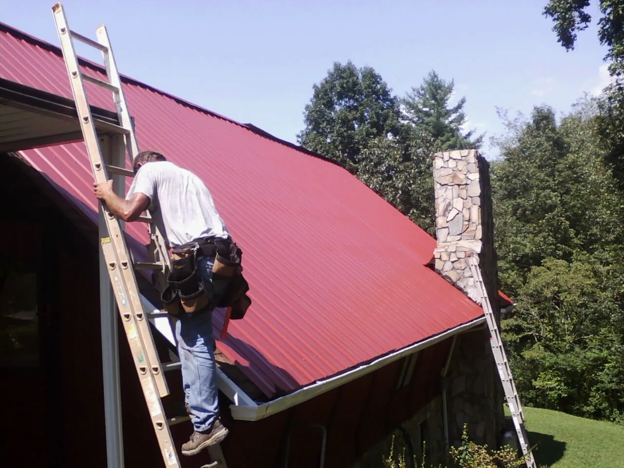 Roofer on a ladder beside a red metal roof and stone chimney — Soffit & Fascia Repair by Roofing Brothers in Creswell